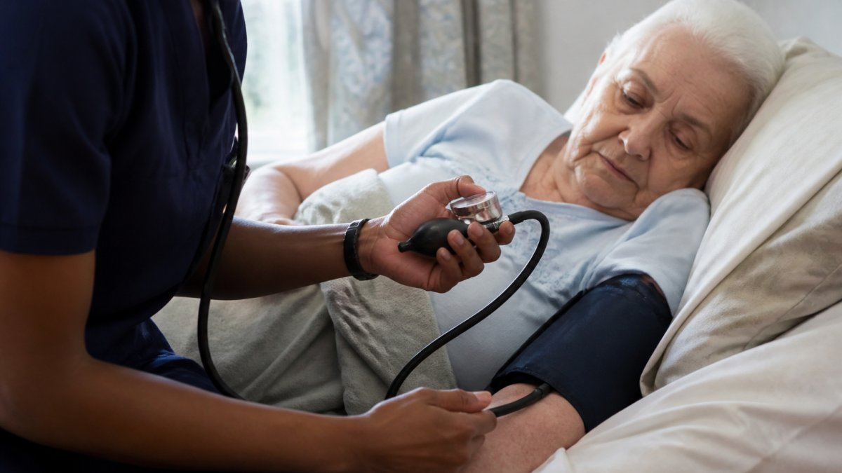 Professional caregiver checking vital signs and assessing a resident's condition with calm, focused demeanor in a residential bedroom setting with soft natural lighting